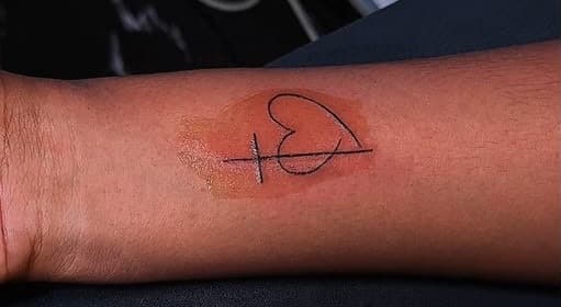 Fine-line tattoo of a heart crossed by a single line on the inner forearm